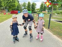 Ein Mann und zwei Mädchen mit Inlinern an den Füßen lächeln in einem Park in die Kamera.