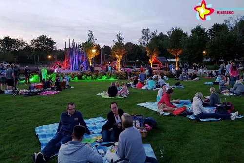 Besucher sitzen auf Picknickdecken, im Hintergrund ist der beleuchtete Spielplatz Schilfwald zu sehen.
