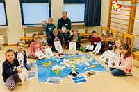 Teilnehmerinnen und Teilnehmer des Kurses "Kleine Klimahelden" sitzen im Halbkreis auf dem Boden. In der Mitte liegen eine große Weltkarte und ein Pinguin.