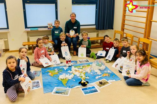 Teilnehmerinnen und Teilnehmer des Kurses "Kleine Klimahelden" sitzen im Halbkreis auf dem Boden. In der Mitte liegen eine große Weltkarte und ein Pinguin.