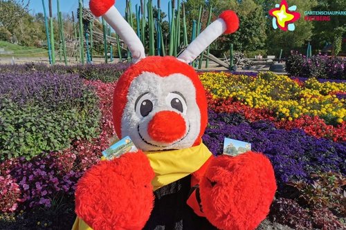 Maskottchen Rieti vor blühenden Blumen, in jeder Hand hält er eine Dauerkarte für den Gartenschaupark.