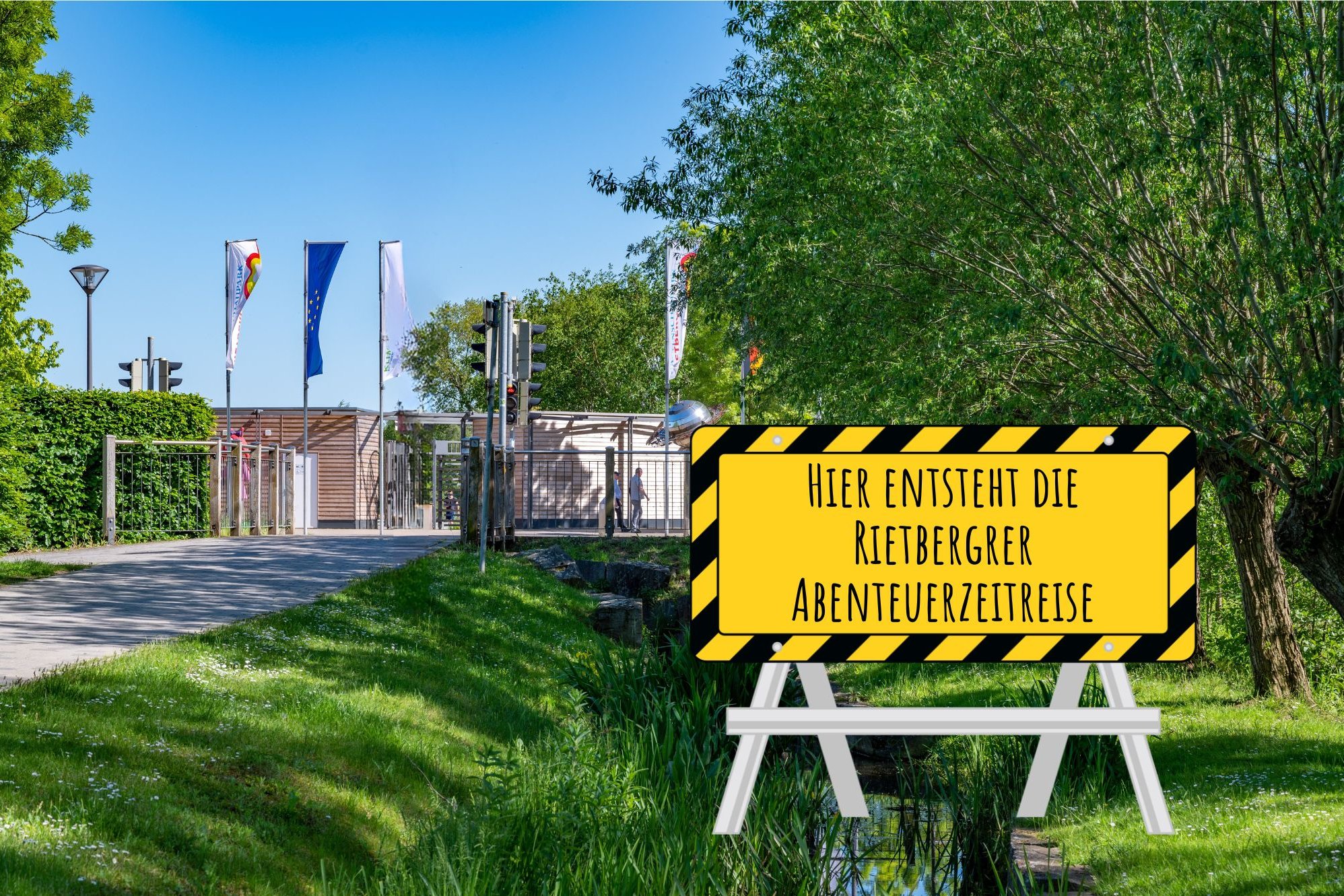 Zu sehen ist der Gartenschaupark-Eingang Mitte aus Richtung Bleiche. Vorne rechts ins Bild montiert ist ein Baustellenschild mit der Aufschrift "Hier entsteht die Rietberger Abenteuerzeitreise".