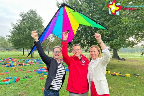 Drei Personen steht auf der großen Wiese im Gartenschaupark und halten einen bunten Lenkdrachen über ihren Köpfen.