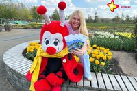 Maskottchen Rieti sitzt im Gartenschaupark auf einer Bank, hinter ihm schaut Bianca Piekatz hervor, die die neuen Flyer zeigt. Im Bild sind auch viele bunte Blümchen zu sehen.