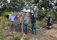 Fünf Frauen und Männer stehen mit Werkzeugen in den Händen nebeneinander in einem Naturgarten.