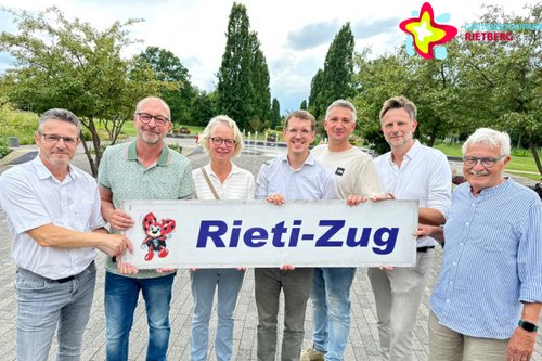 Sieben Frauen und Männer stehen in einem Park und halten gemeinsam ein Schild mit der Aufschrift "Rieti-Zug".