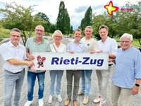 Sieben Frauen und Männer stehen in einem Park und halten gemeinsam ein Schild mit der Aufschrift "Rieti-Zug".