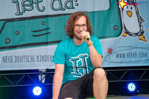 herrH hockt auf der Bühne, das Mikro vor dem Mund.