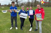 Spendenlauf-Organisatoren und Park-Geschäftsführer stehen auf einer Wiese im Park und halten eine Starterklappe, einen Ball, einen Zettel mit dem Wort "Start", eine Urkunde und das Plakat zum Spendenlauf in Händen.