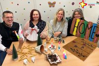 Geschäftsführer und Grünes-Klassenzimmer-Mitarbeiterinnen sitzen hinter einem Tisch, auf dem jede Menge Gebasteltes liegt: kleine Gipsfiguren, ein Engel aus Papier, Tannenbäume etc. Iris Peter hält eine selbst gestaltete Fußmatte mit der Aufschrift "HOME" hoch.