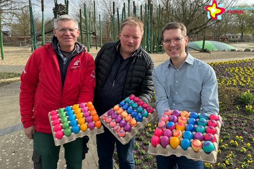 Chefgärtner Marco Rüschkamp, Thomas Determeyer und Johannes Wiethoff (von links) stehen vor dem Spielplatz Schilfwald und halten jeweils eine Palette mit bunten Ostereiern in die Kamera.