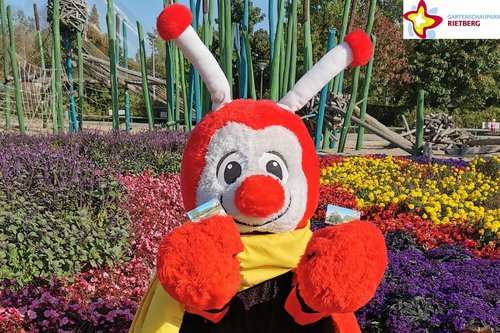 Maskottchen Rieti vor Blumenbeet und Spielplatz mit Dauerkarten.