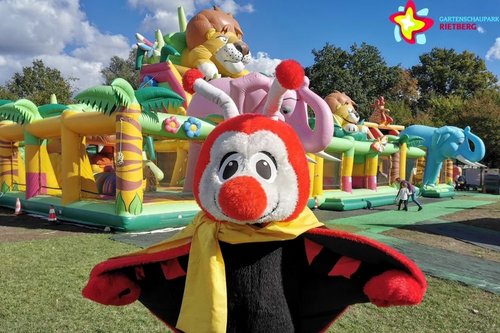 Maskottchen Rieti steht vor der Hüpfburg Dschungelland mit zwei großen aufgeblasenen Elefantenköpfen. Im Hintergrund ist der Kopf der Löwen-Hüpfburg zu sehen.