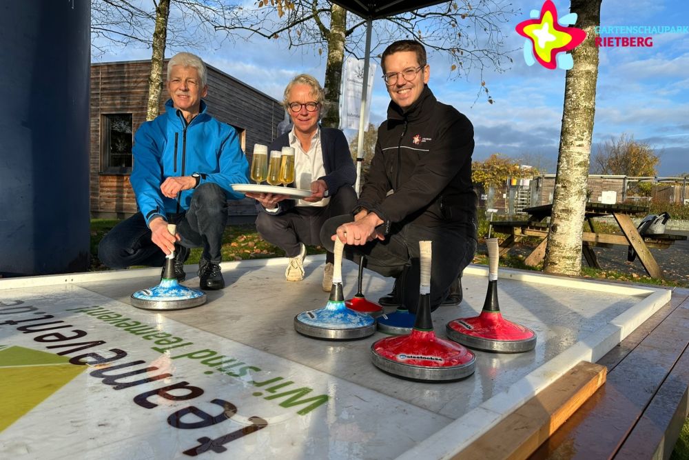 Rainer Kornfeld, Anja Rodenbeck und Johannes Wiethoff hocken auf der Bahn, im Vordergrund sind mehrere Eisstöcke zu sehen. Anja Rodenbeck hält ein Tablett mit gefüllten Biergläsern in der Hand.