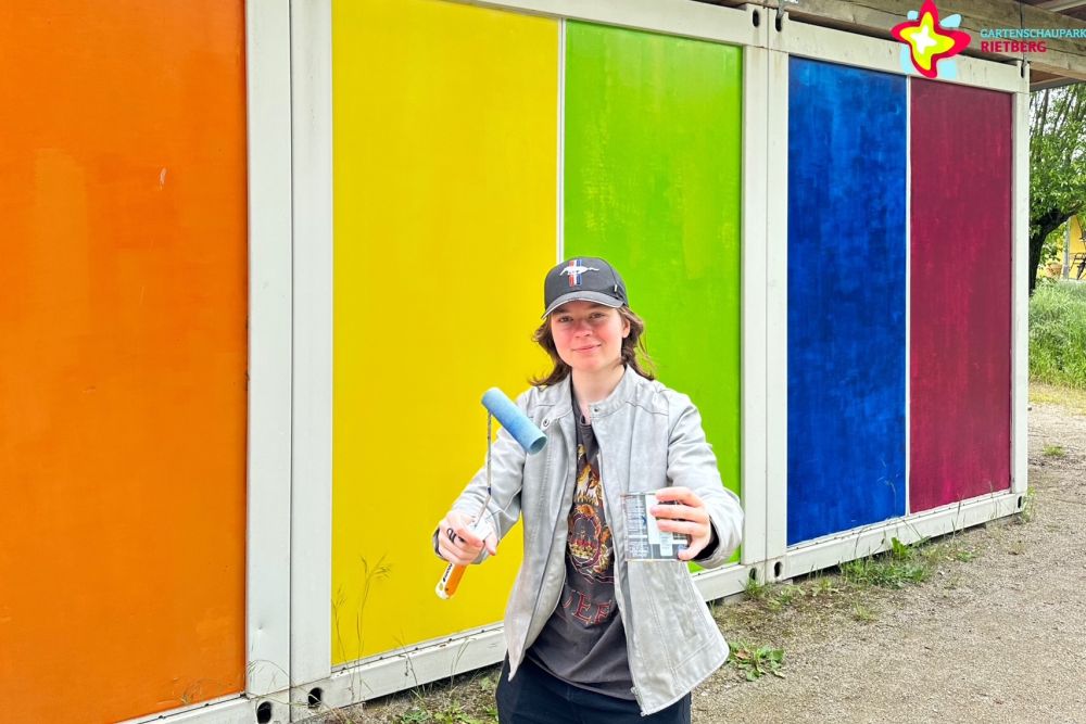 Leah Schmelz mit Farbtopf und Farbrolle in der Hand vor dem bunten Container des Grünen Klassenzimmers.