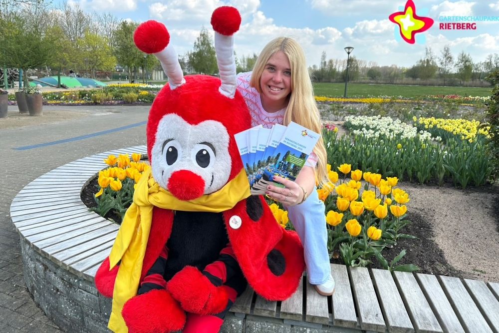 Maskottchen Rieti sitzt im Gartenschaupark auf einer Bank, hinter ihm schaut Bianca Piekatz hervor, die die neuen Flyer zeigt. Im Bild sind auch viele bunte Blümchen zu sehen.