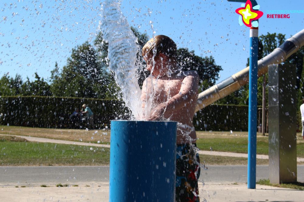 Ein Junge auf dem Wasserspielplatz hinter einer Wasserfontäne.