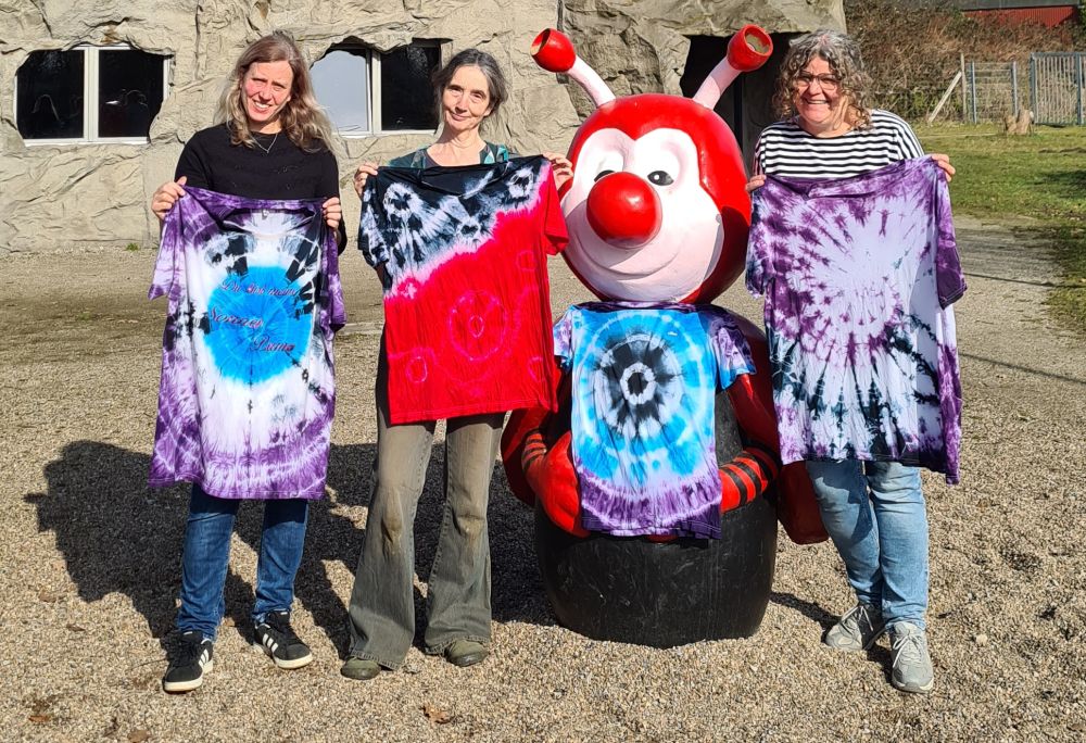 Zu sehen sind die drei Mitarbeiterinnen aus dem Grünen Klassenzimmer, die gebatikte T-Shirts in Händen halten. Sie stehen rechts und links neben einer Rieti-Figur, die ein gebatiktes T-Shirt trägt.