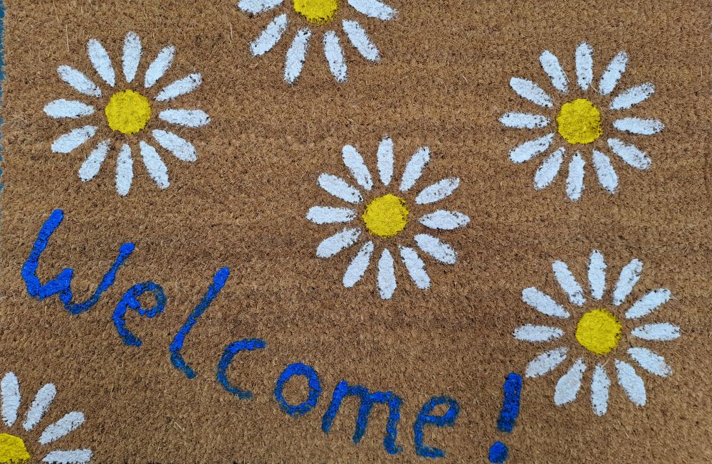 Eine Fußmatte mit mehreren Blümchen, zu lesen ist das Wort "Welcome".