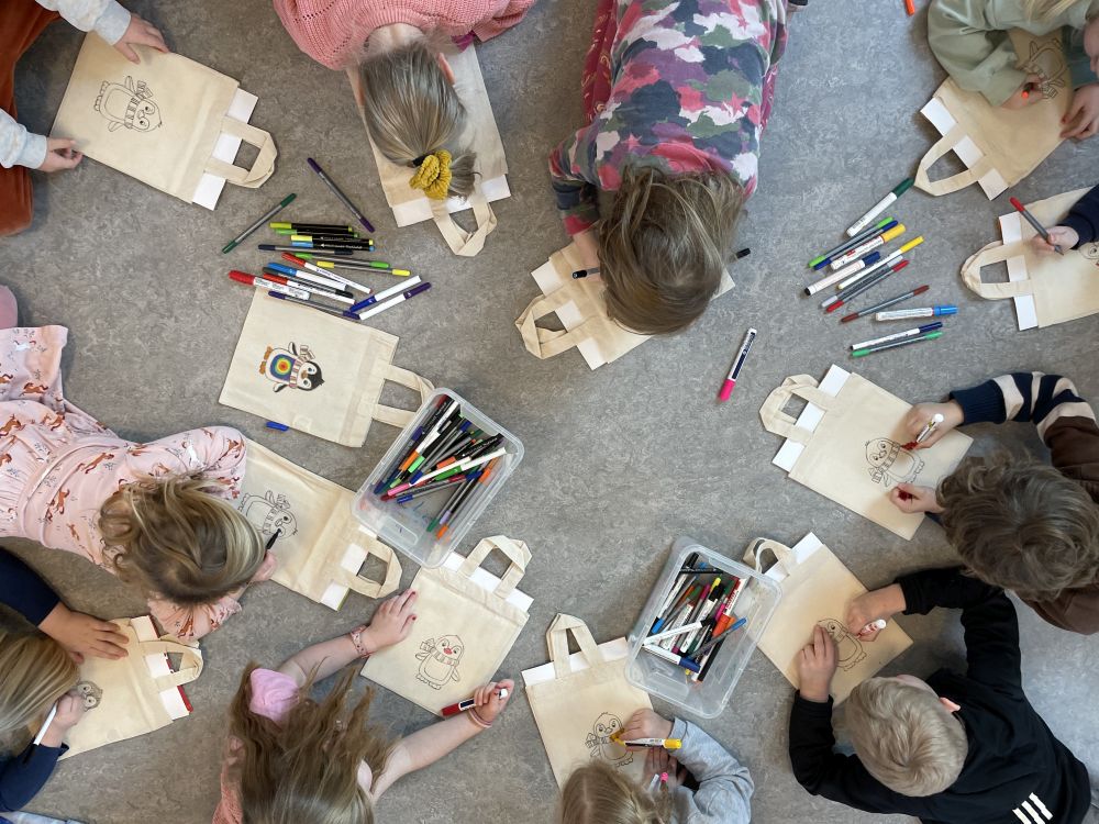 Ansicht von oben: Kinder liegen auf dem Boden und bemalten Beutel mit Pinguinen.
