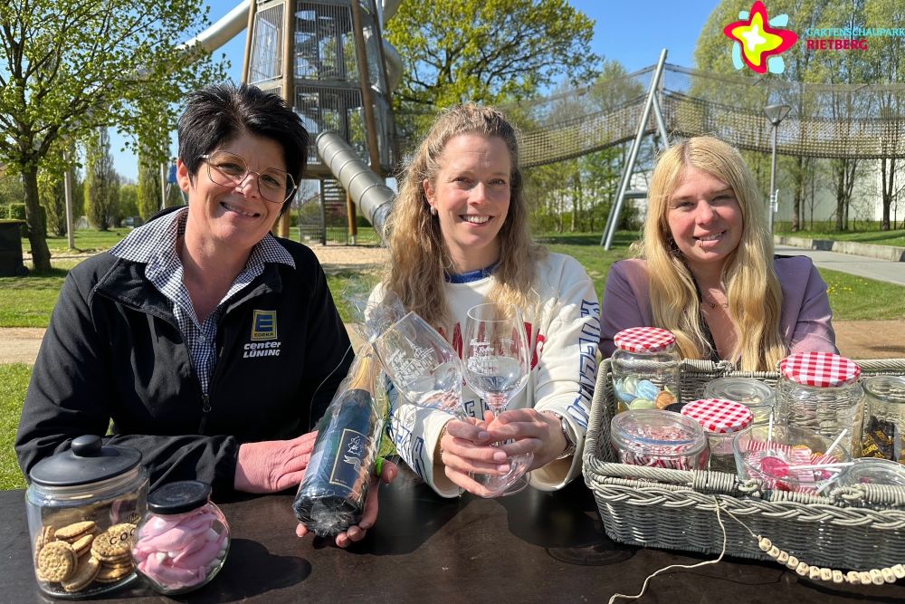 Die Gartenschaupark-Mitarbeiterinnen Bianca Piekatz und Laura Hunke sitzen mit Nicole Börnemeier vom E-Center Lüning an einem Tisch im Gartenschaupark und halten eine Sektflasche, Gläser und andere Picknick-Utensilien in die Kamera. Im Hintergrund ist der große Rutschenturm zu sehen.