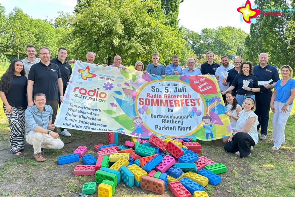 Teilnehmer der Entdeckerzone, Organisatoren und Veranstalter mit einem großen Radio-Gütersloh-Sommerfest-Banner. Im Vordergrund sind die Großbausteine von der Picknickwiese im Gartenschaupark zu sehen.