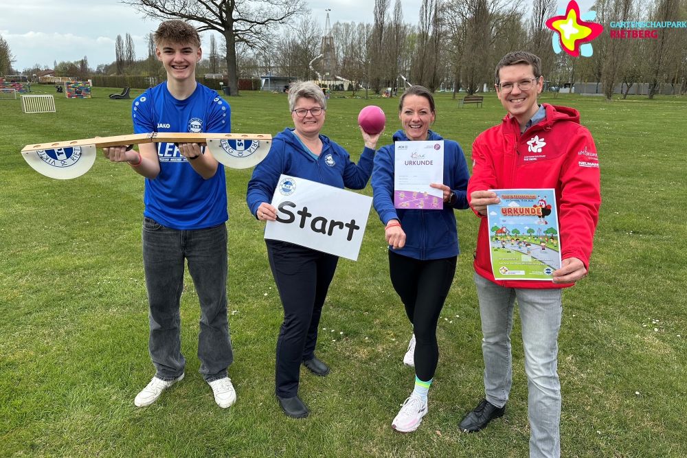Spendenlauf-Organisatoren und Park-Geschäftsführer stehen auf einer Wiese im Park und halten eine Starterklappe, einen Ball, einen Zettel mit dem Wort "Start", eine Urkunde und das Plakat zum Spendenlauf in Händen.